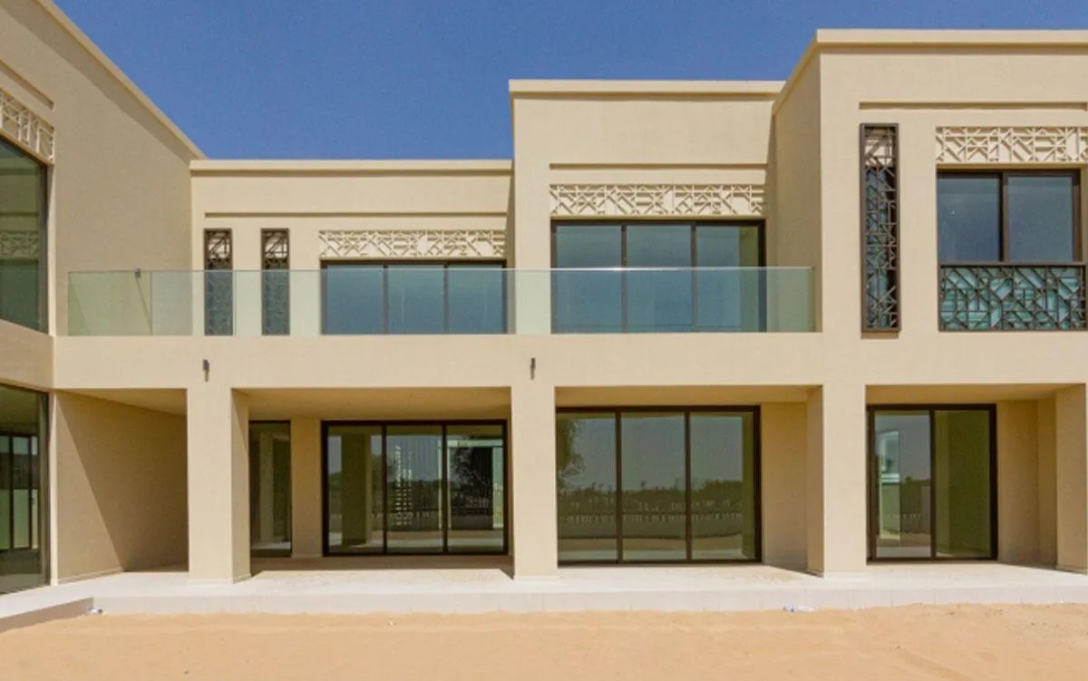 Villa elevation in warm stone render with pierced geometric parapet screens, a glass balustrade to the first floor terrace and full-height sliding glazing below.