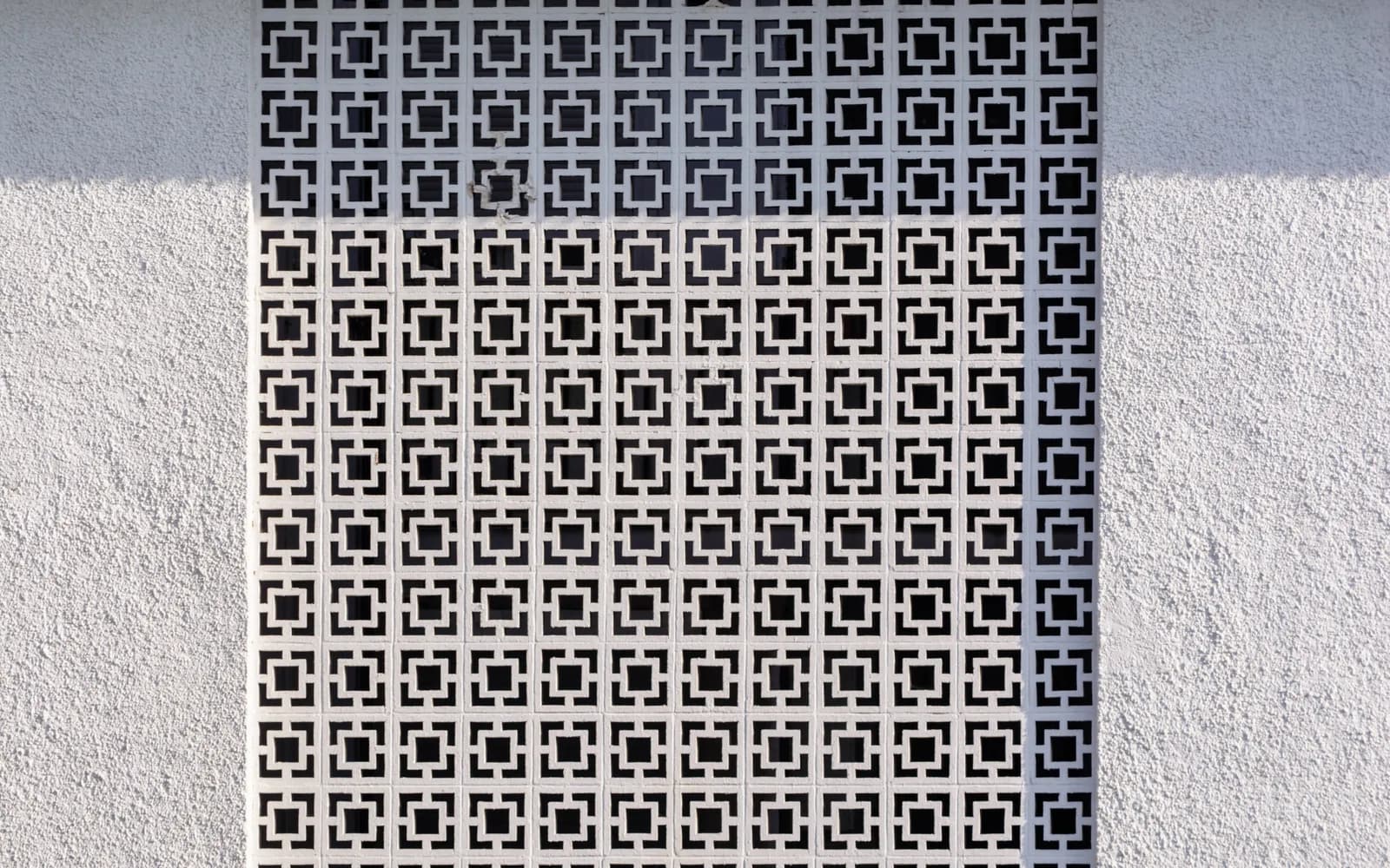 Pierced concrete jali screen in a geometric repeating pattern, set into a textured white render wall in raking sunlight.