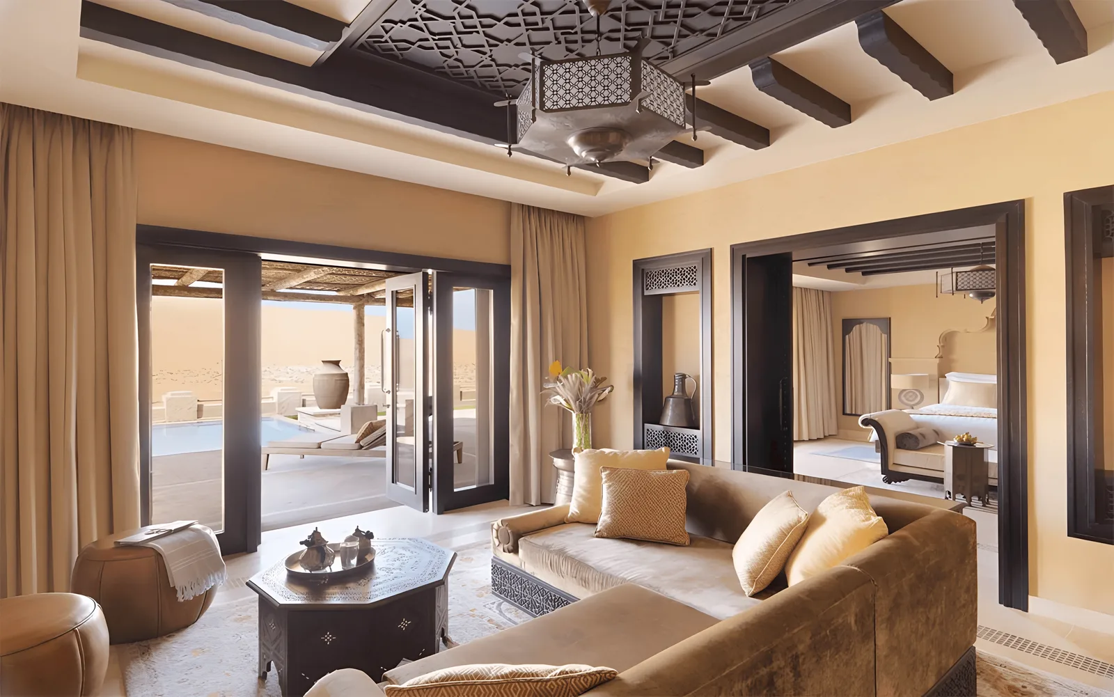 Living room with a carved dark-timber ceiling, exposed beams, a pierced metal pendant lantern, mashrabiya niches and polished sand-coloured plaster, opening through folding doors to a pool terrace.