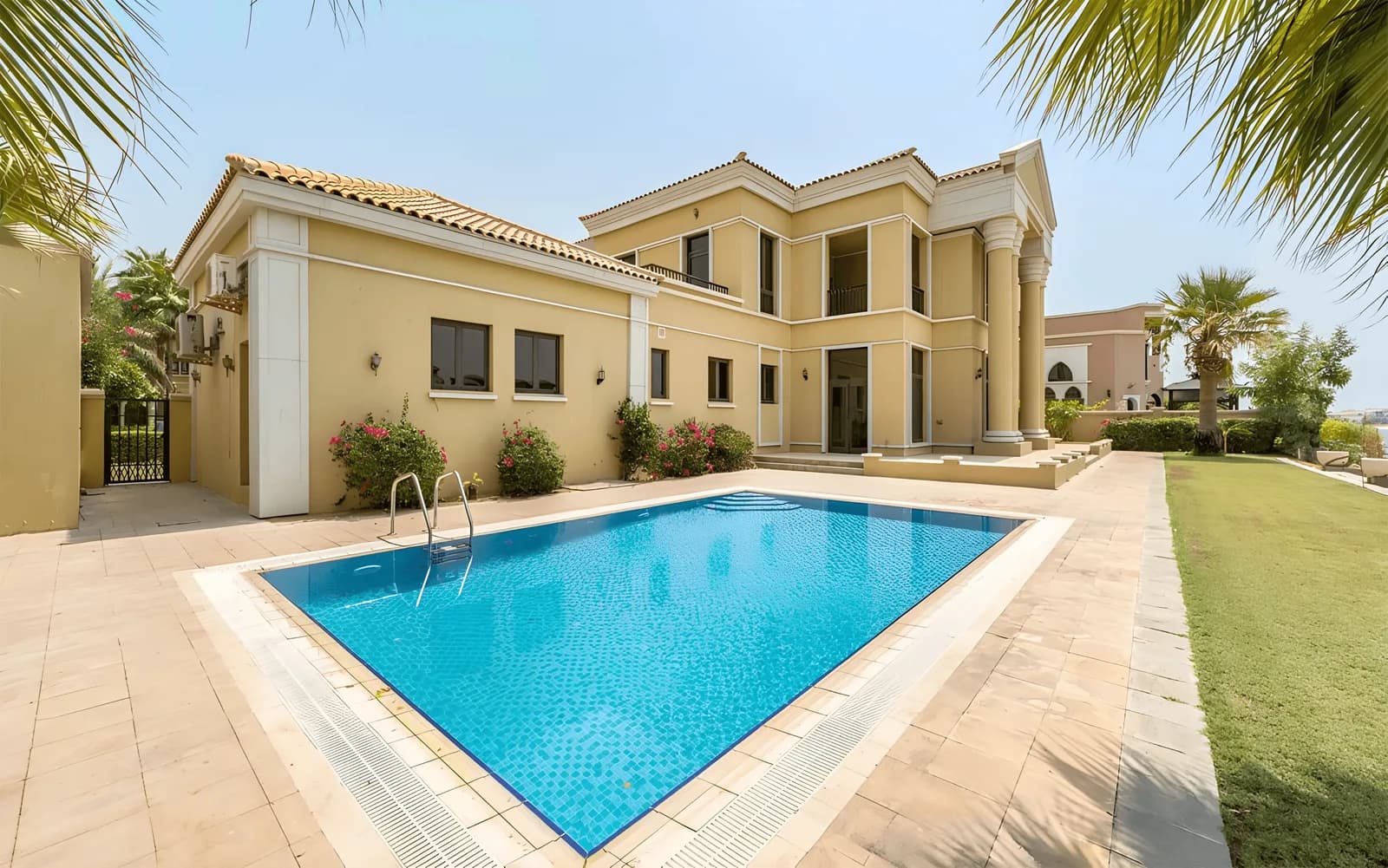 Villa with a columned portico and a tiled swimming pool set in a paved terrace with flowering shrubs and lawn.